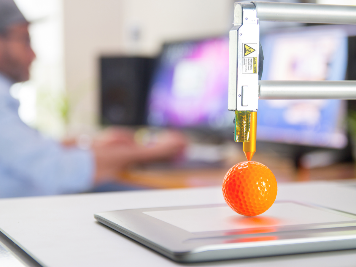 3D printer arm finishes printing neon orange golf ball, which sits on platform on the table. Man in the background sits looking at two computer screens on a separate desk.
