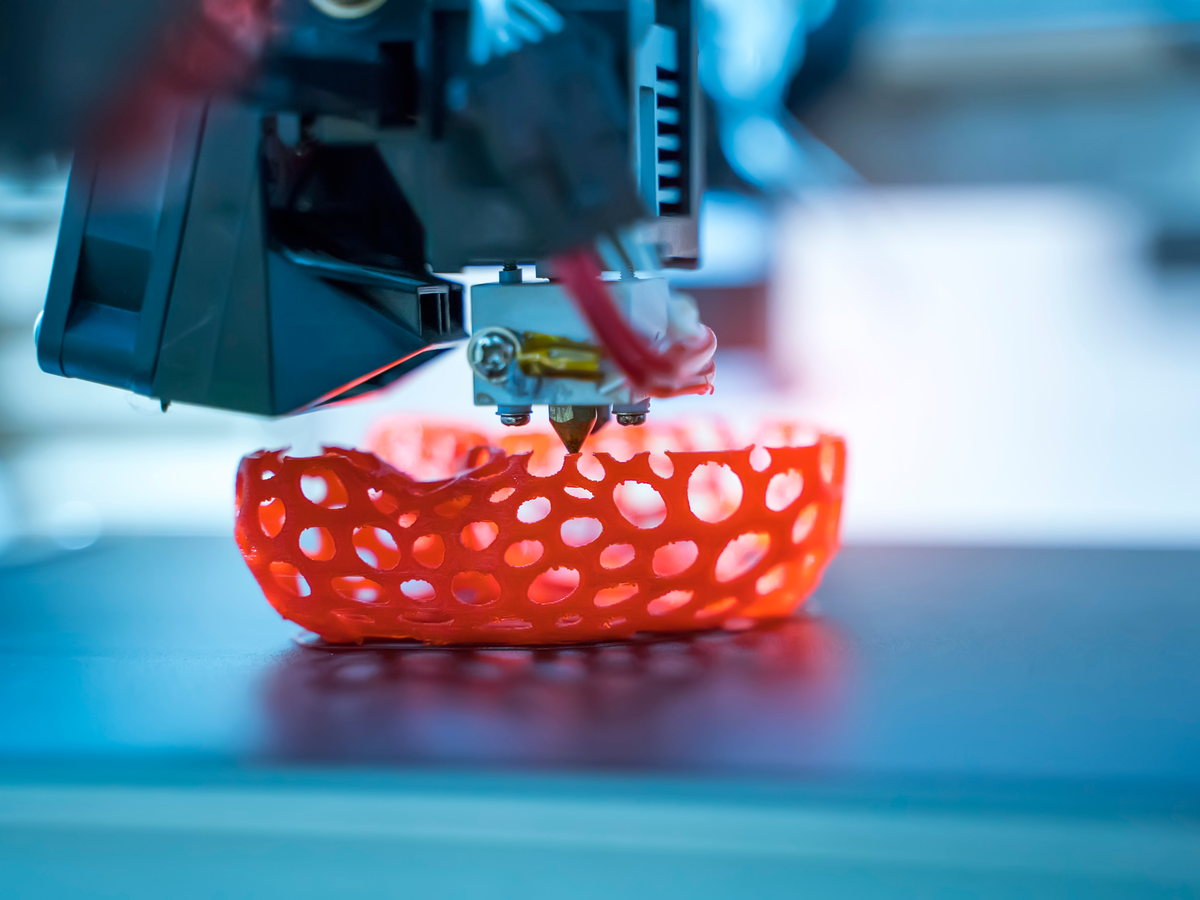3D printing machine in the process of printing a red, net-like object which sits on a platform.