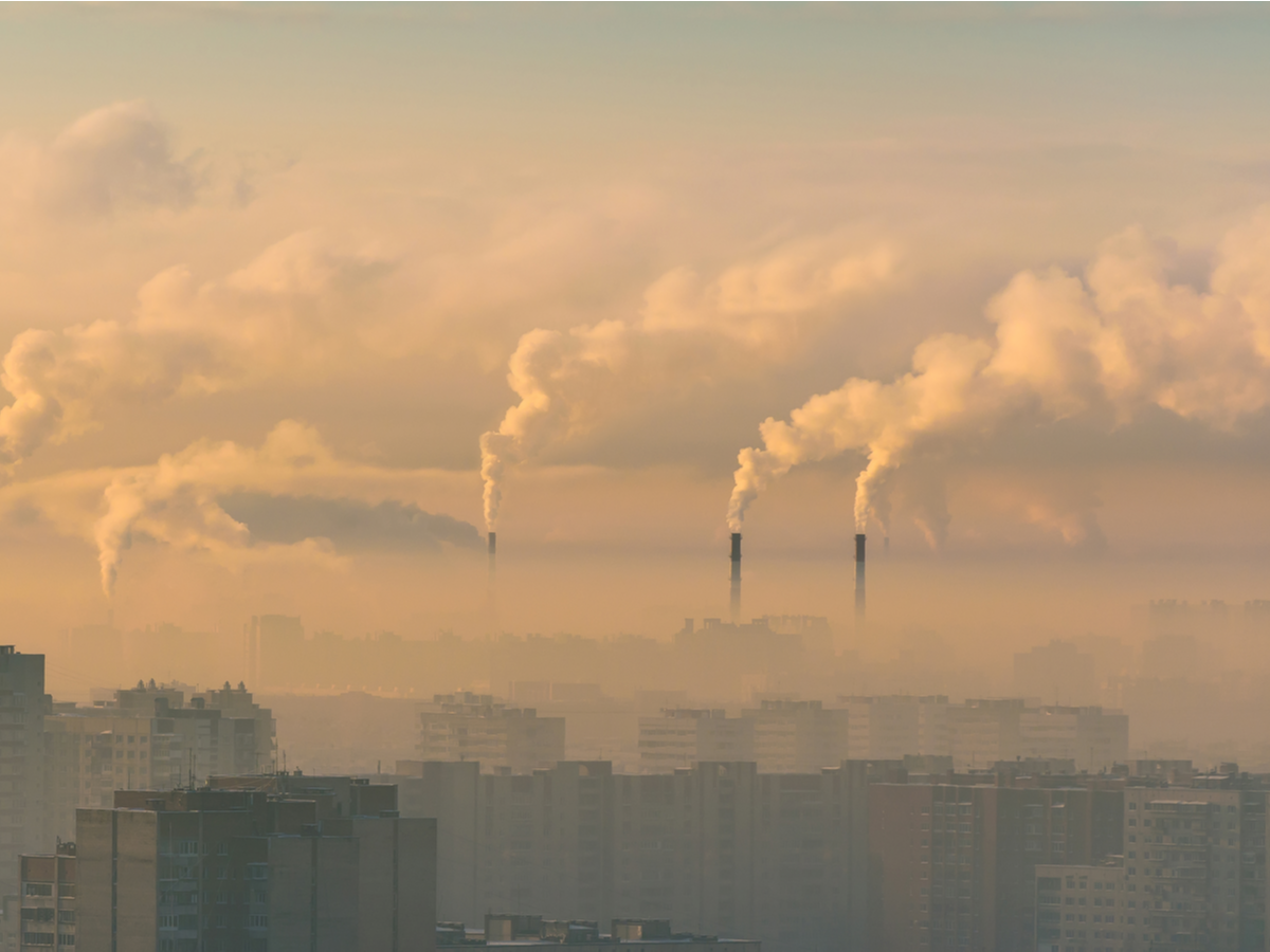 City skyline partially obscured by smog, which is thicker towards the background. Three smokestacks can be seen above the layer of smog, pouring smoke.
