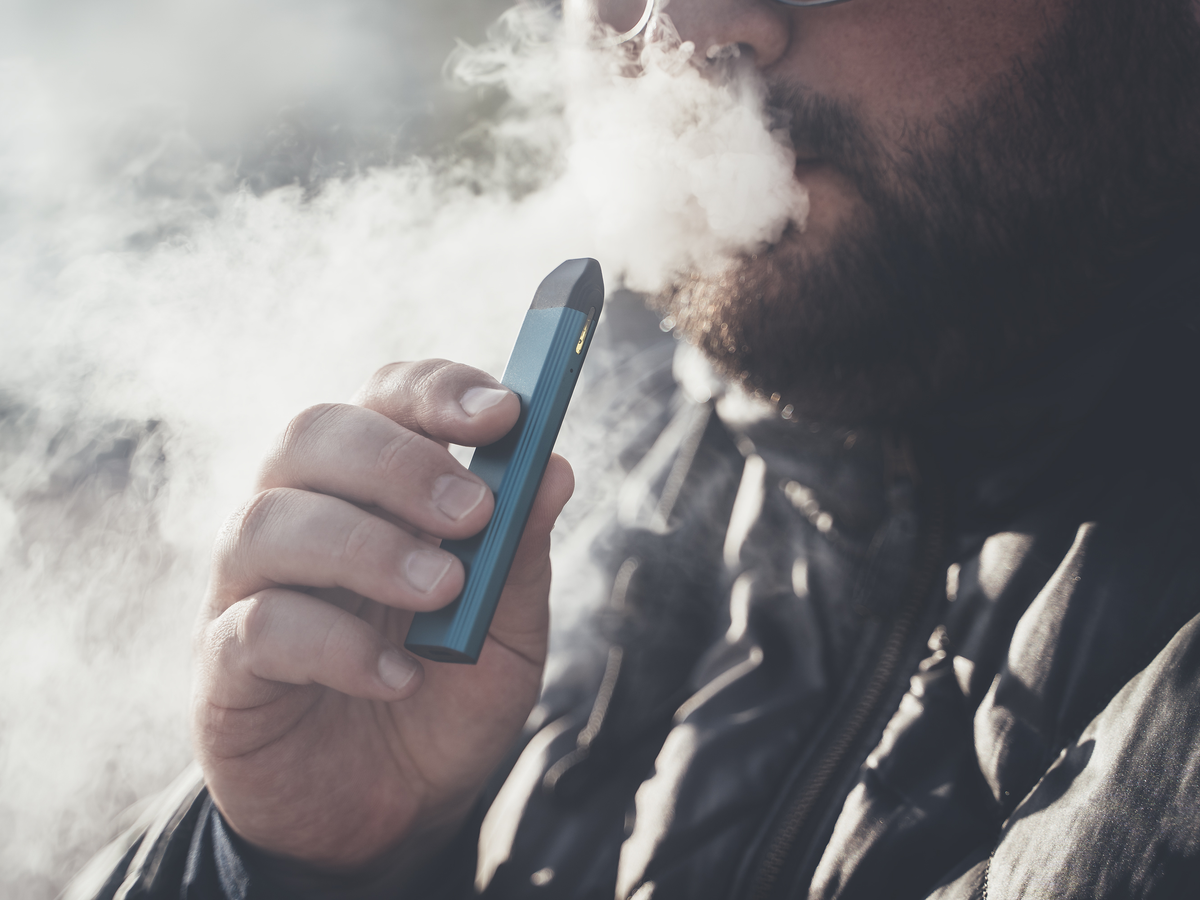 Close up of man with beard and moustache holding vaporizer near mouth and exhalling cloud of smoke.