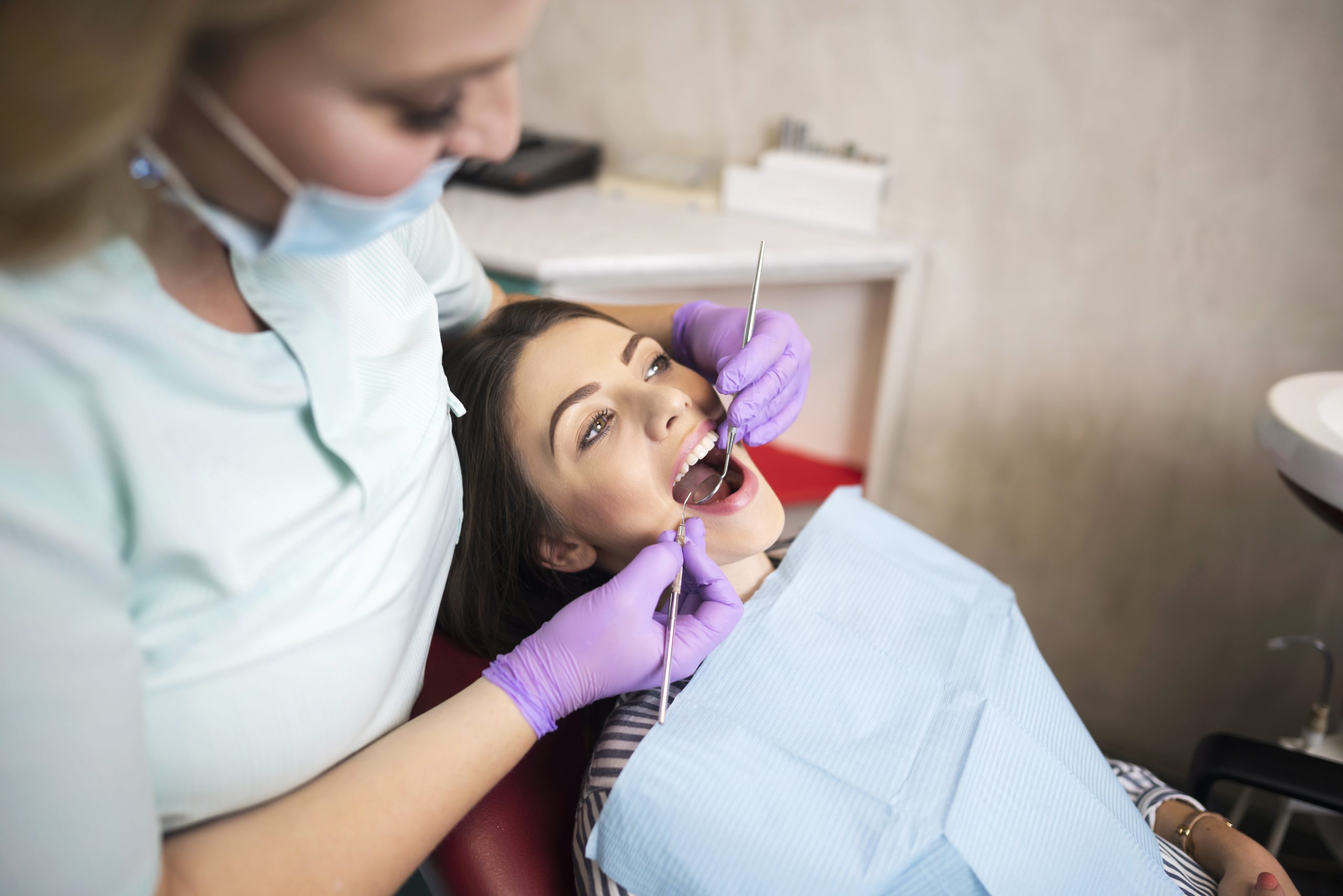 Female receiving dental checkup from woman wearing purple gloves and holding dentistry tools.