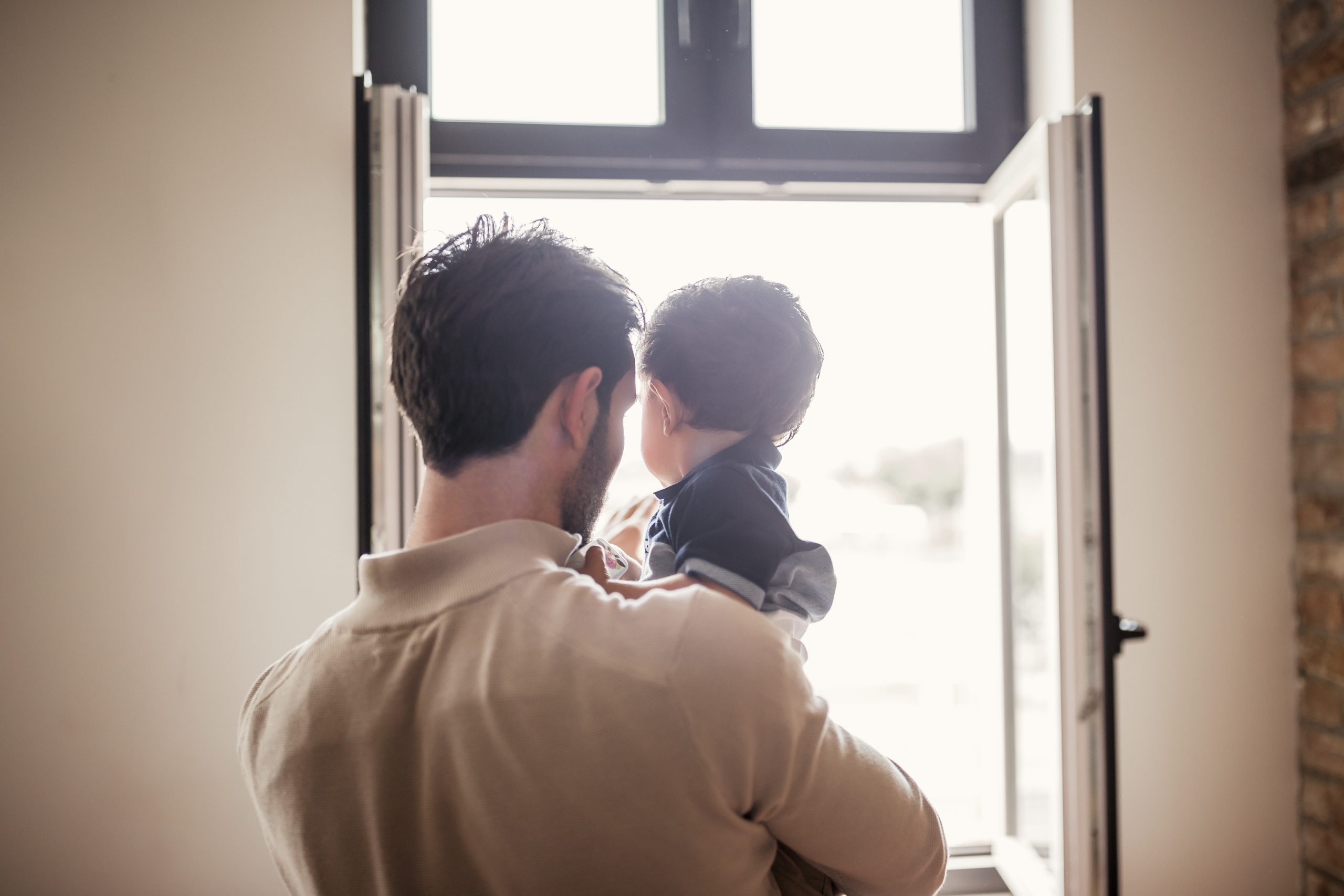 Man holding baby boy standing near open window.