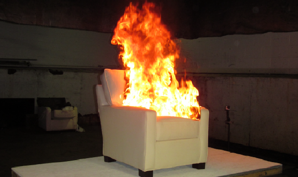 White lounge chair sits on industrial platform. Large flames rise from the seat cushion and obscure the back of the chair